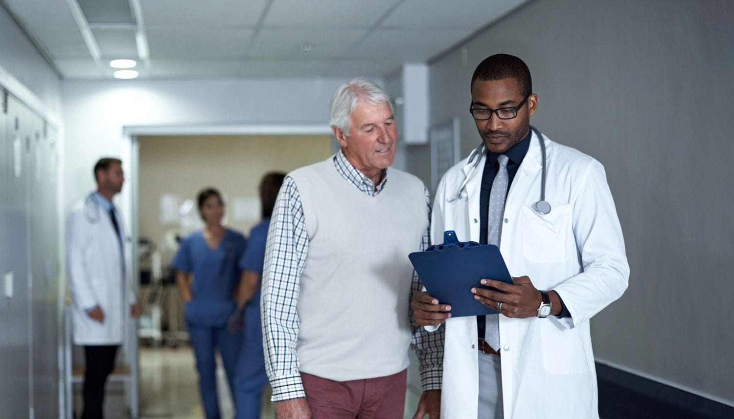 Doctor talking to an older patient while holding a clipboard and standing in a hospital corridor