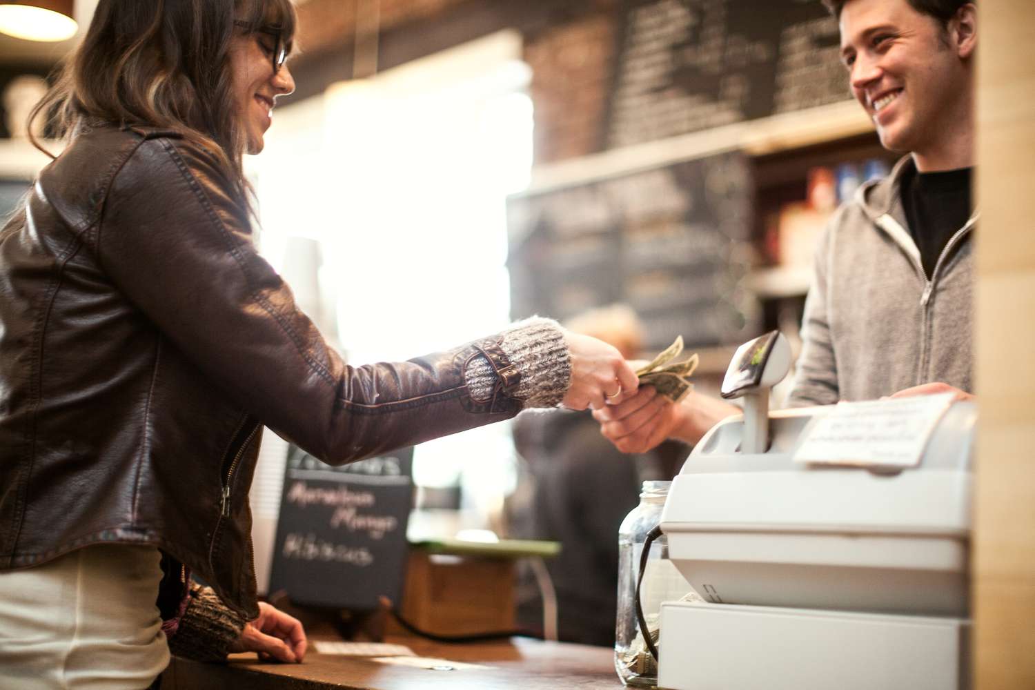 A woman is paying at a coffee shop. The cashier is a young man. They are smiling at each other.