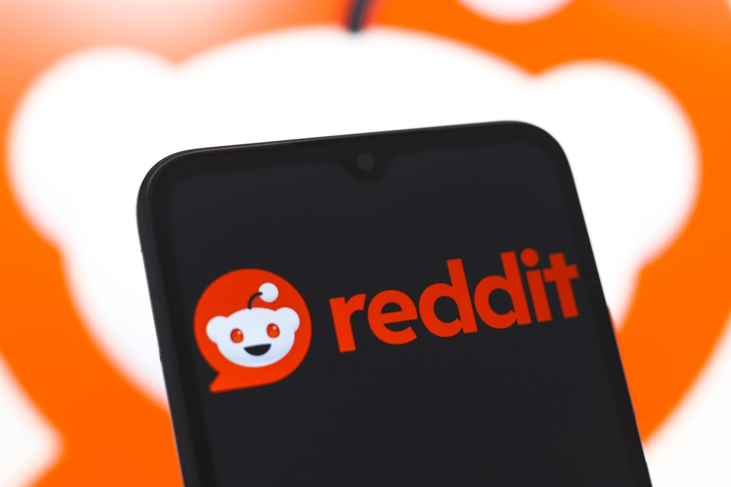 The orange Reddit logo is seen on a cell phone screen in front of a larger version of the logo in the background.