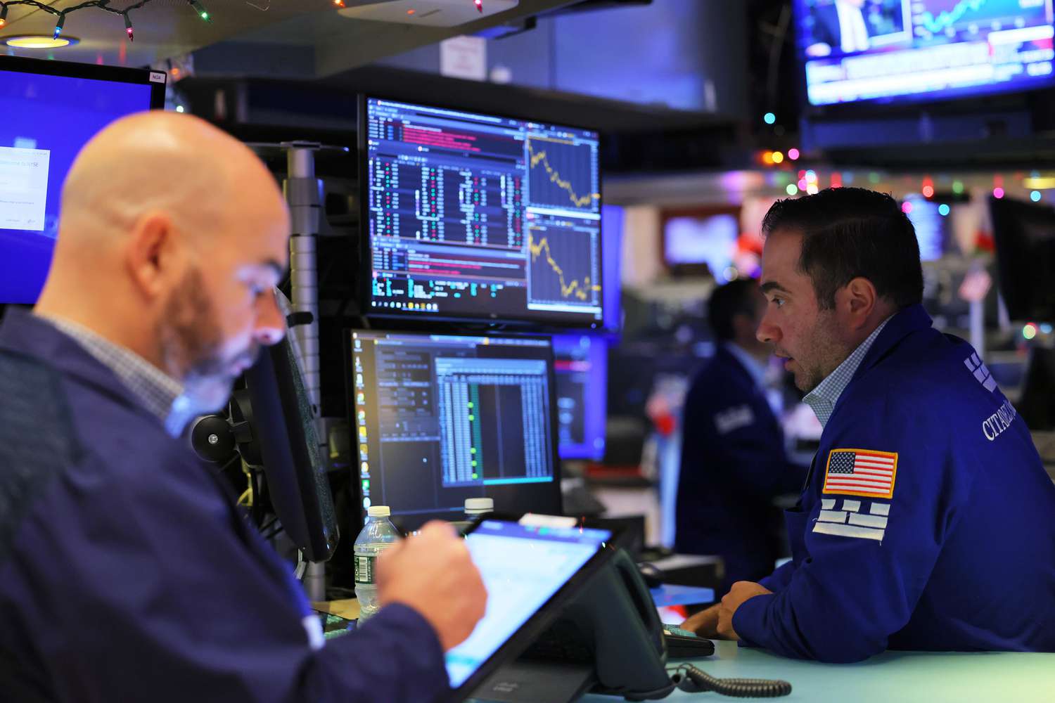 Two traders work on the floor of the New York Stock Exchange in front of stock charts