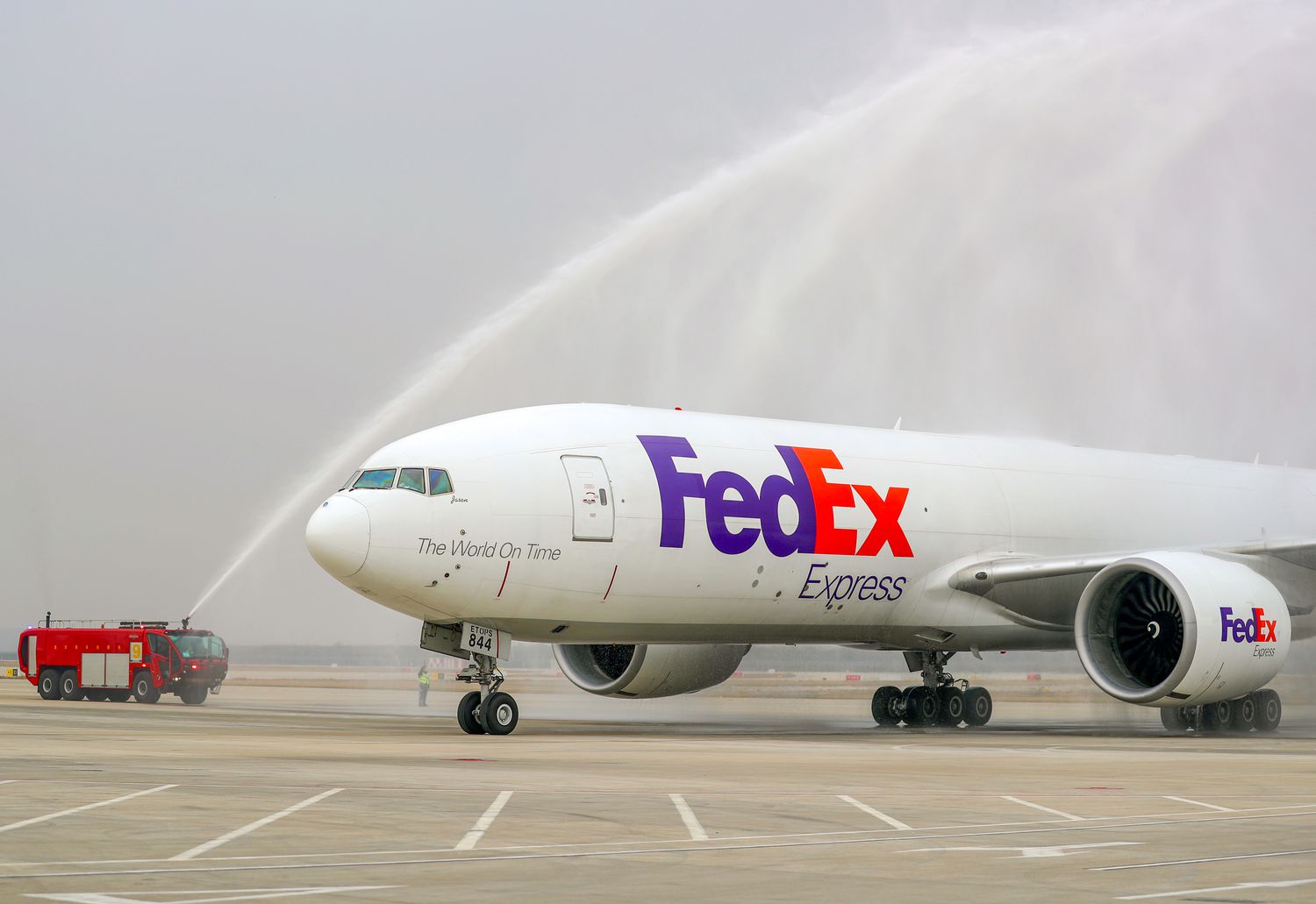 A red fire truck sprays water in the direction of a white FedEx Express airplane.