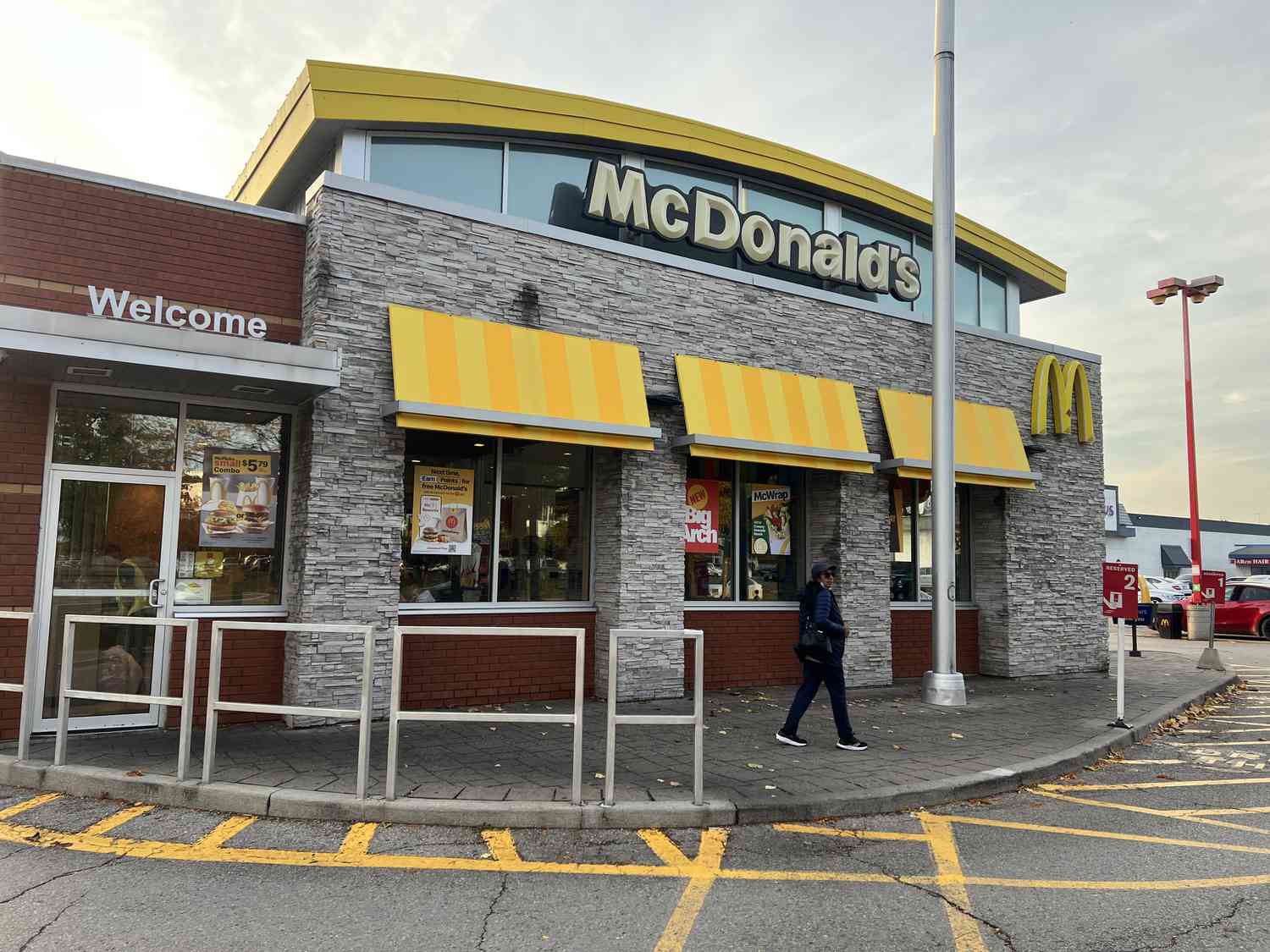 A person is seen walking outside a gray, brown, and yellow McDonald's restaurant with a blue-gray sky behind it.