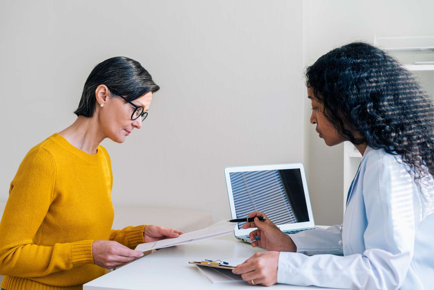 A doctor with dark hair talks with a patient wearing glasses about a recent insurance claim.