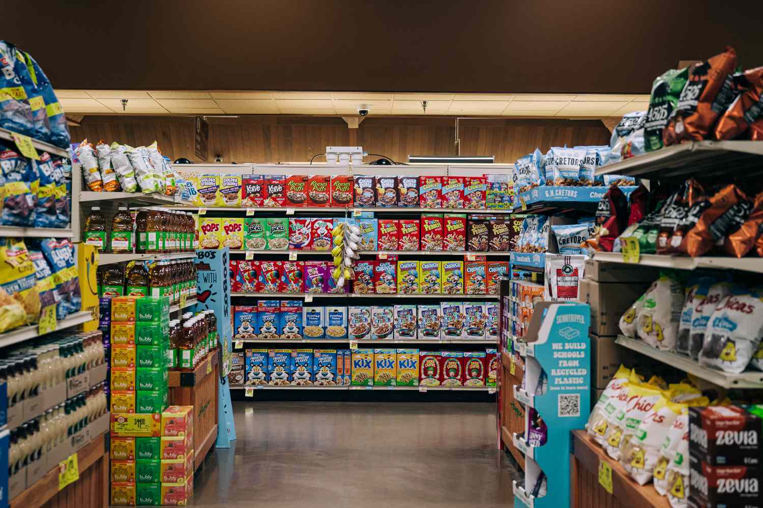 A grocery aisle is seen in a store, with boxes of cereal and bags of chips on shelves.