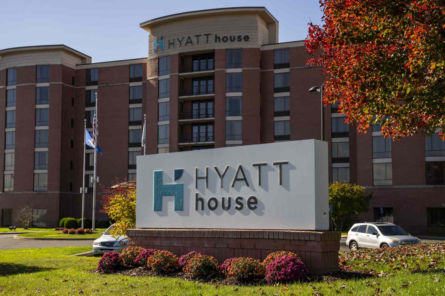 A Hyatt House sign in front of a hotel, with grass, bushes, a tree, and two cars around the sign