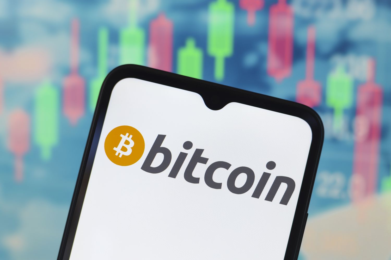 The Bitcoin logo is seen on a phone screen with the green and red bars of a stock chart in the background.