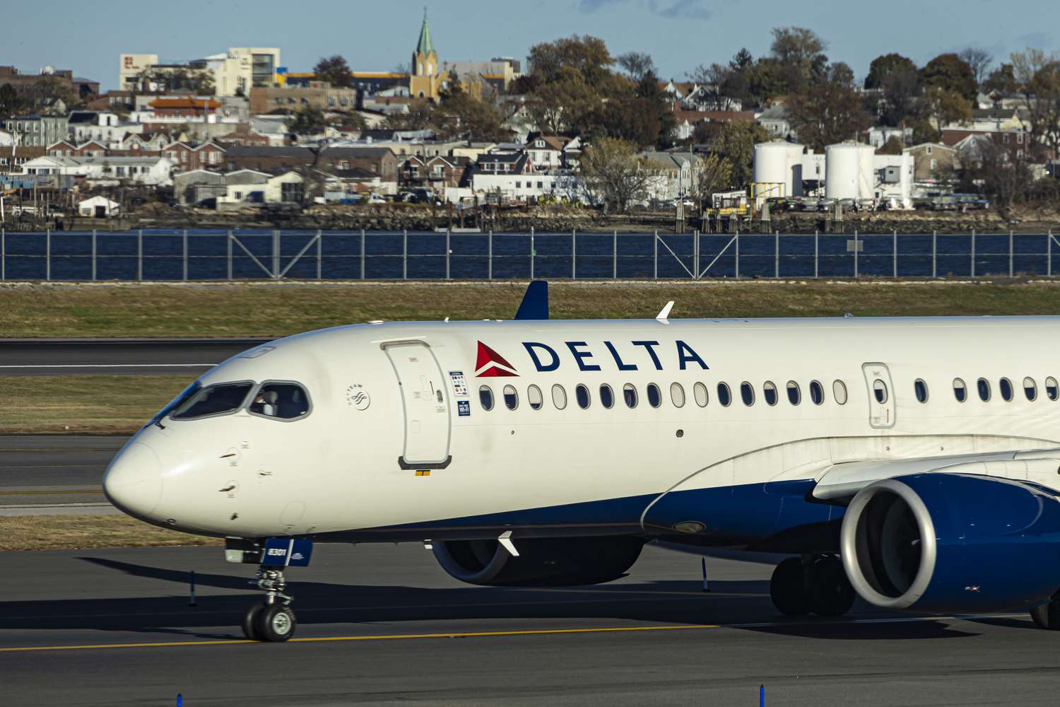 A white Delta Air Lines plane is seen on an airport runway with a city in the background.