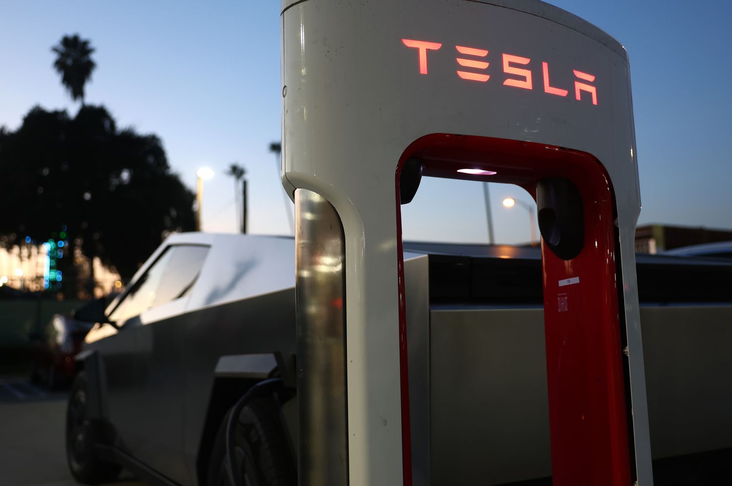 A white and red Tesla charger is seen in front of a Tesla Cybertruck and a sunset.